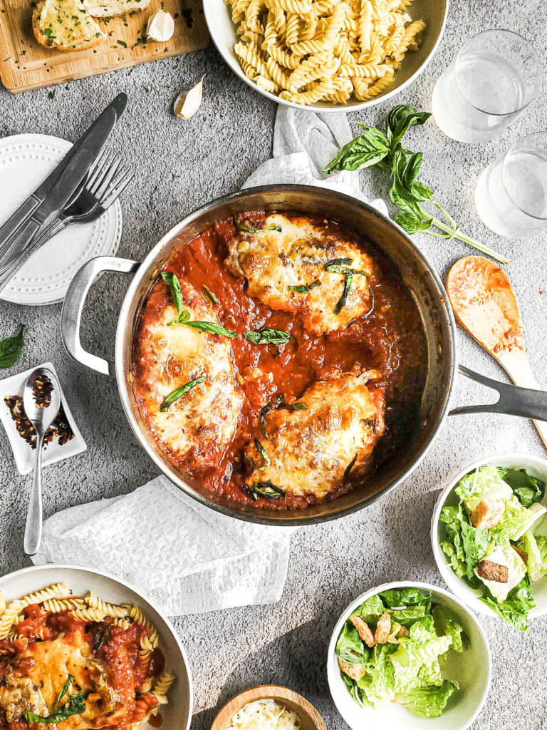 Overhead shot of chicken parmesan in a pan with a salad and bread on the side