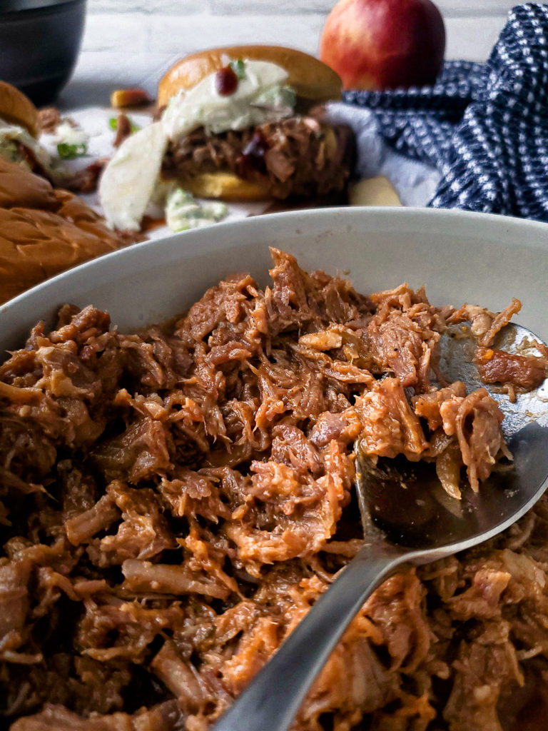 Close up of pulled pork in a bowl with a pulled pork sandwich in the background