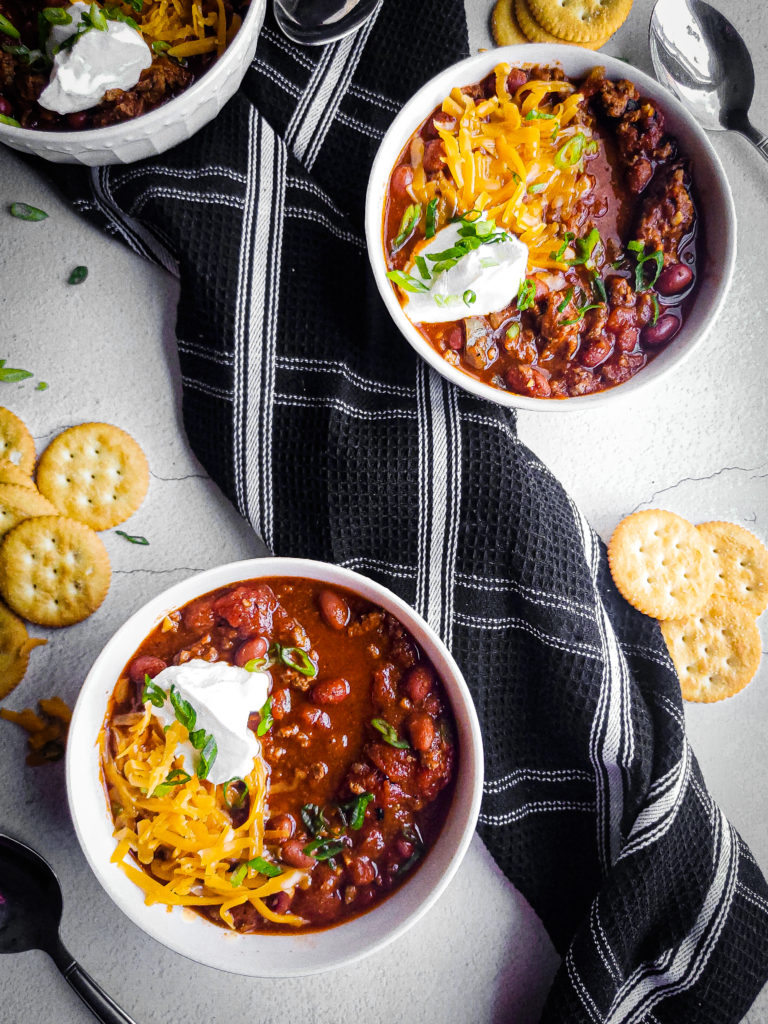 Overhead shot of 3 bowls of beef chili with shredded cheese, sour cream, and crackers